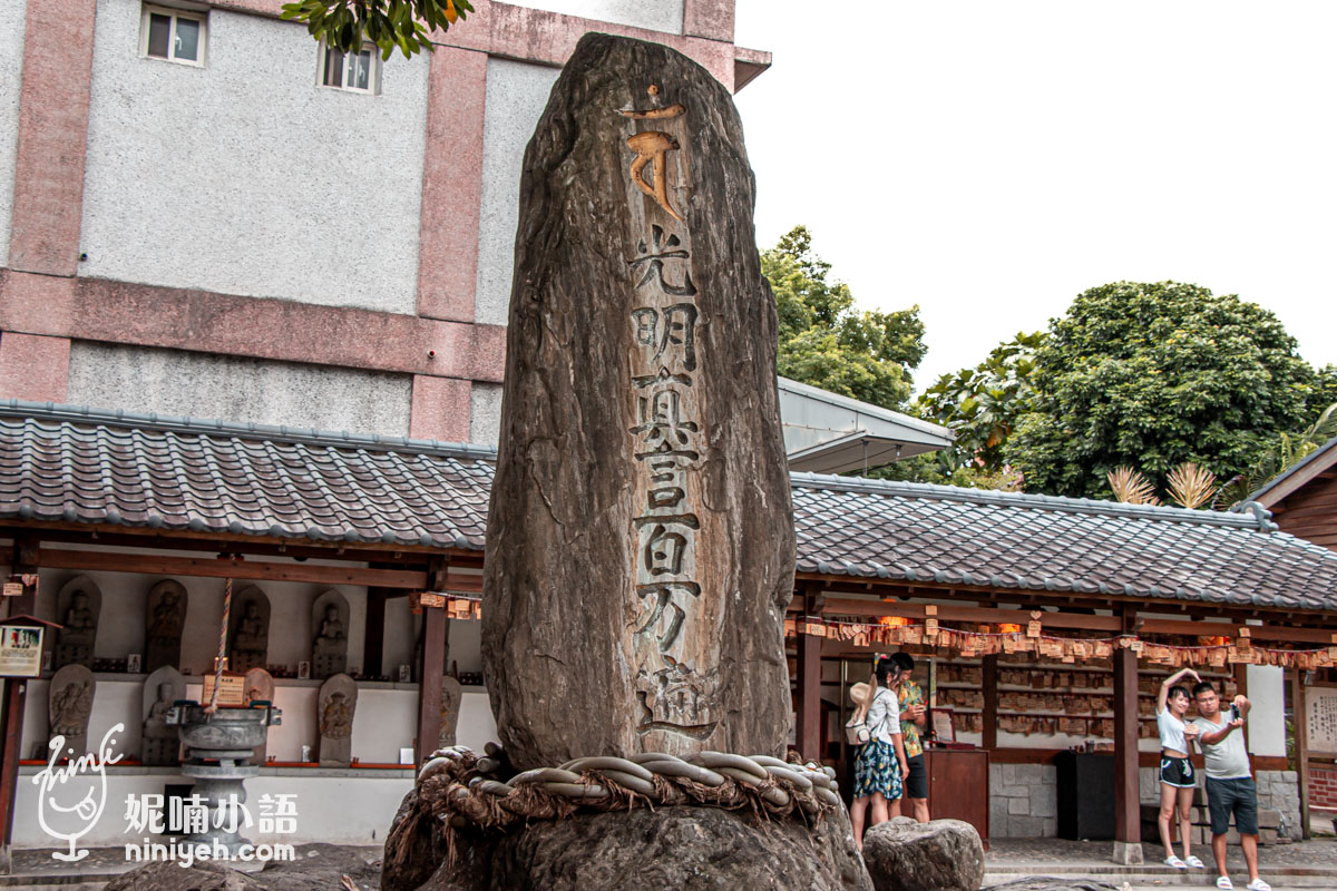 吉安慶修院庭園中央由木柵欄圍繞的光明真言百萬遍石碑全景，後方為陳列石佛的黑瓦長廊與遊客