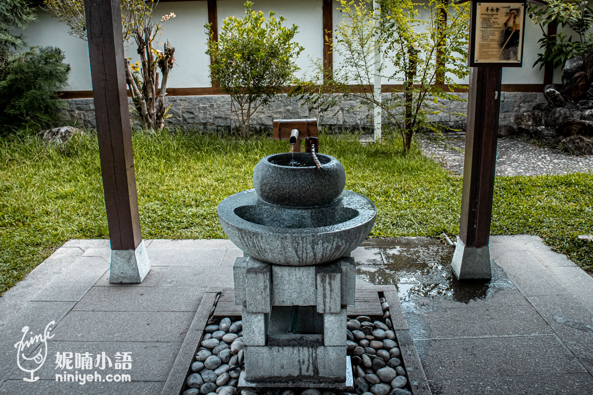 吉安慶修院手水舍的雙層灰藍色石雕水槽，頂部竹管緩緩流出清澈流水，下方鋪滿灰色鵝卵石