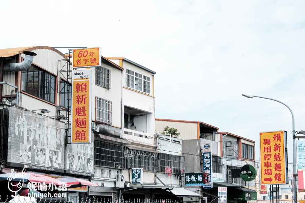 賴新魁麵館的店面外觀，顯示60年老字號的招牌，並標示創立於1945年，旁邊還有專用停車場的指示牌，位於苗栗三義的街道上。