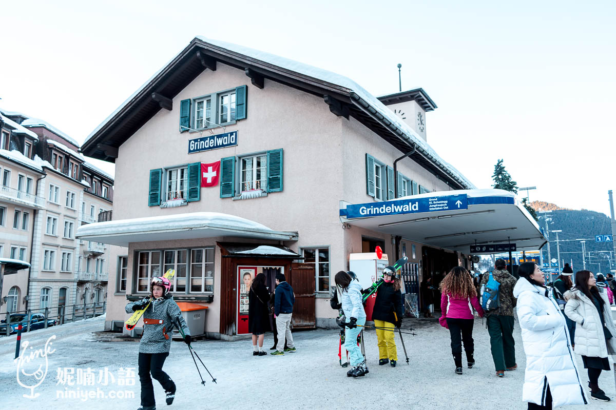 瑞士格林德瓦（Grindelwald）火車站的冬日景象，背景是一棟典雅的建築，外牆掛著瑞士國旗，帶有典型的阿爾卑斯風情。火車站前聚集著穿著滑雪裝備的遊客，手持滑雪板和雪杖，準備前往滑雪場，顯示這裡是熱門的滑雪勝地。周圍被雪覆蓋的環境與遊客的活力，呈現出格林德瓦作為滑雪小鎮的熱鬧氛圍。