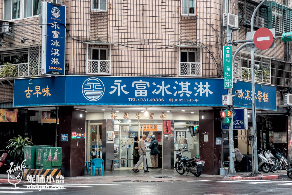 台北萬華區的永富冰淇淋店面，這家擁有超過70年歷史的老店自1945年創立以來，堅持提供古早味冰品。店面位於街角，顯眼的藍色招牌上清楚地寫著「永富冰淇淋」和「古早味」，以及創立年份，帶給人一種懷舊的氛圍。店門口簡單乾淨，旁邊還設置了幾張椅子，方便顧客稍作停留享用冰品。