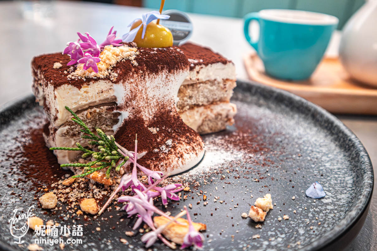 義式提拉米蘇甜點切片，灑滿可可粉並淋上鮮奶油，點綴金色巧克力球、紫色花朵與餅乾碎屑，背景有藍色茶杯與茶壺，擺盤精美。