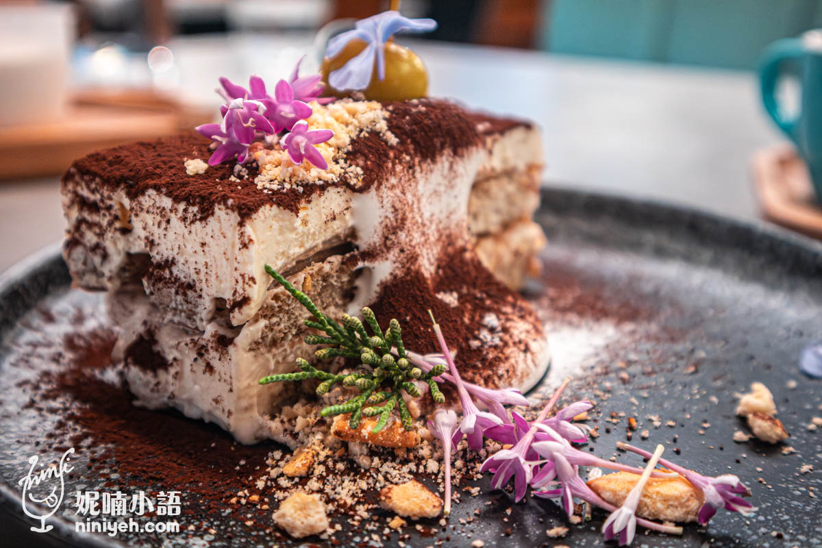提拉米蘇切片擺盤,表面灑滿可可粉並點綴金色巧克力球、紫色花朵與餅乾碎屑,旁邊配有鮮奶油與香草葉,視覺精緻如藝術品。