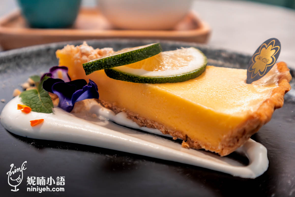 檸檬派切片特寫,派面放有兩片新鮮檸檬片與薄荷葉,搭配紫色可食用花與白色鮮奶油醬,擺盤精緻且色彩鮮明。