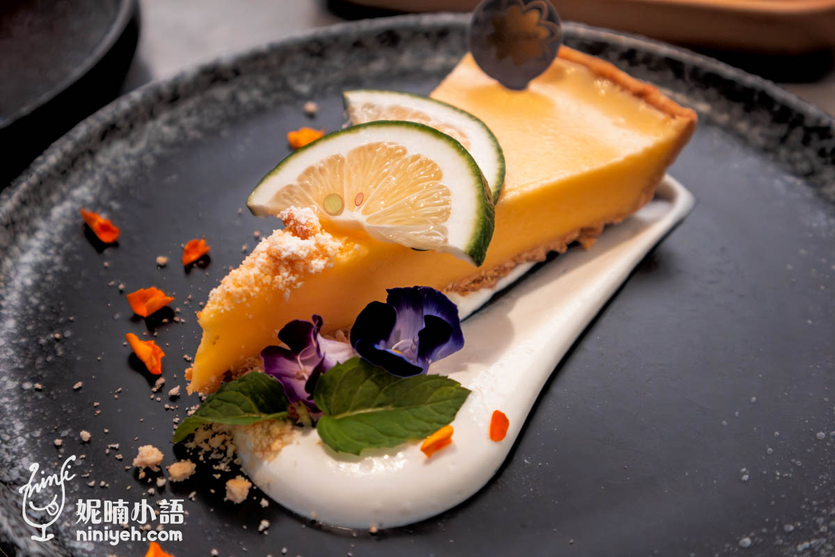 檸檬派切片擺放在黑色圓盤上,派面裝飾兩片新鮮檸檬片與薄荷葉,搭配紫色可食用花與白色鮮奶油醬,點綴橙色花瓣,擺盤精緻。