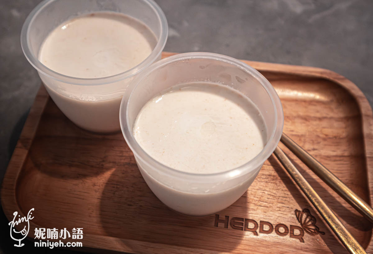 兩杯透明塑膠杯裝的花生奶酪放在木盤上,奶酪表面光滑細緻,旁邊擺有金色湯匙,木盤刻有HERDOR字樣。