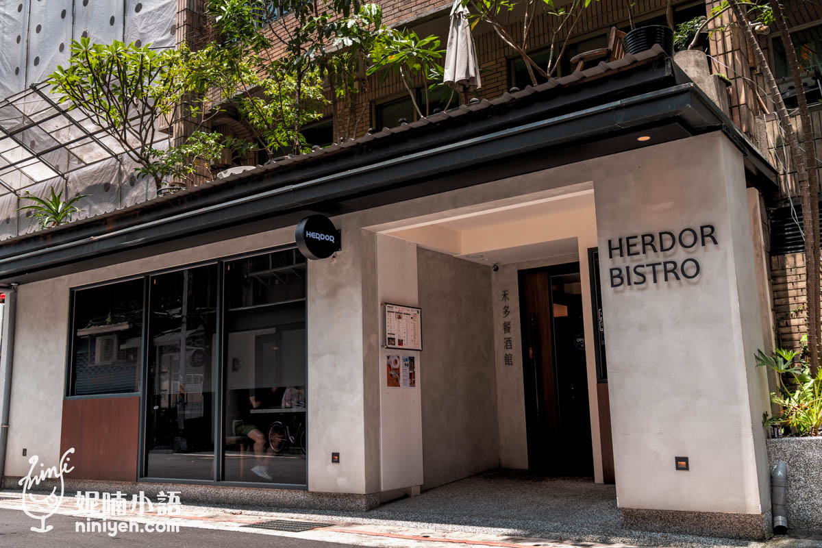 禾多餐酒館(HERDOR BISTRO)外觀,灰色水泥牆搭配黑色窗框與招牌,店門口有植物點綴,位於台北市街道旁