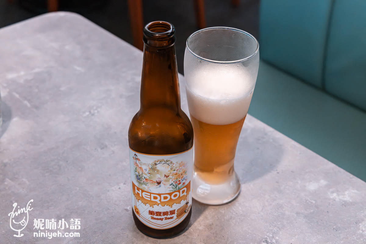 HERDOR蜂蜜啤酒與冰鎮啤酒杯並排放在桌上，啤酒杯中充滿金黃色酒液與綿密泡沫，瓶身標籤圖案精緻，呈現清爽風格。