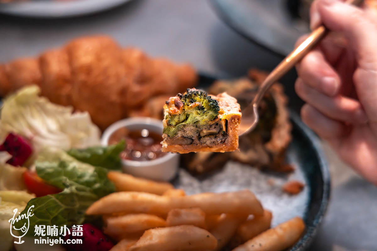 特寫拍攝叉子夾起一塊西蘭花野菇鹹派,內餡飽滿包含香菇、蘑菇與融化起司,背景可見可頌、薯條與生菜沙拉,色澤誘人。