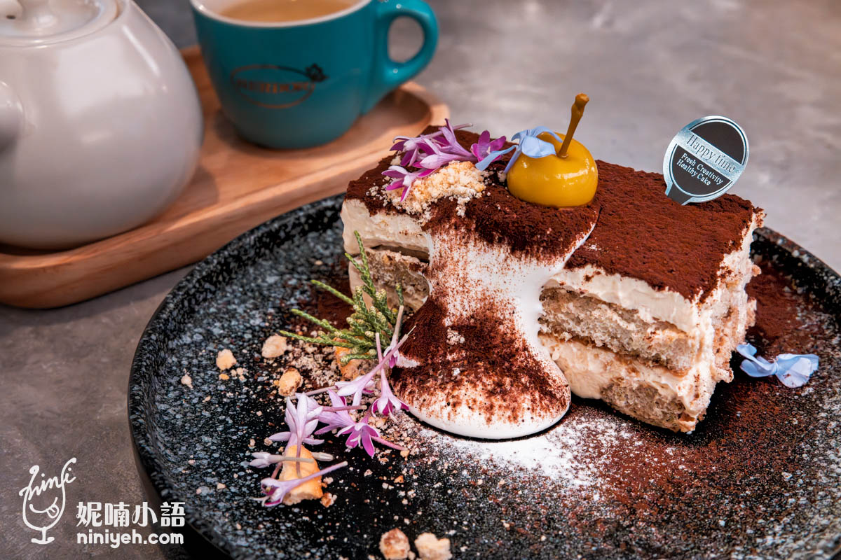 精緻提拉米蘇甜點擺盤,表面撒滿可可粉並點綴金色巧克力裝飾與小花,旁邊配有鮮奶油,背景可見一杯茶飲。
