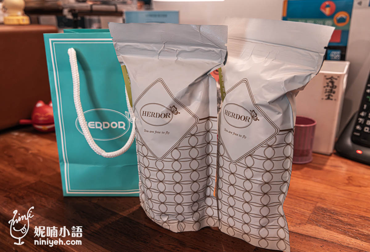 禾多餐酒館外帶茶包,兩袋HERDOR罐裝罐蓋荔香茶擺放在木桌上,旁邊有一個藍綠色HERDOR紙袋,背景為溫馨室內環境。
