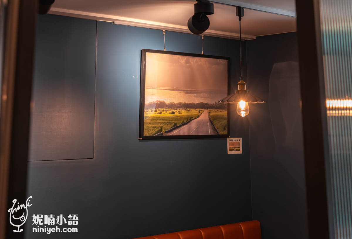 禾多餐酒館牆面掛有鄉村道路與田園風景照片,旁邊懸掛一盞復古吊燈,燈光暖黃,營造溫馨氛圍,下方可見部分橙色皮質長椅座位。