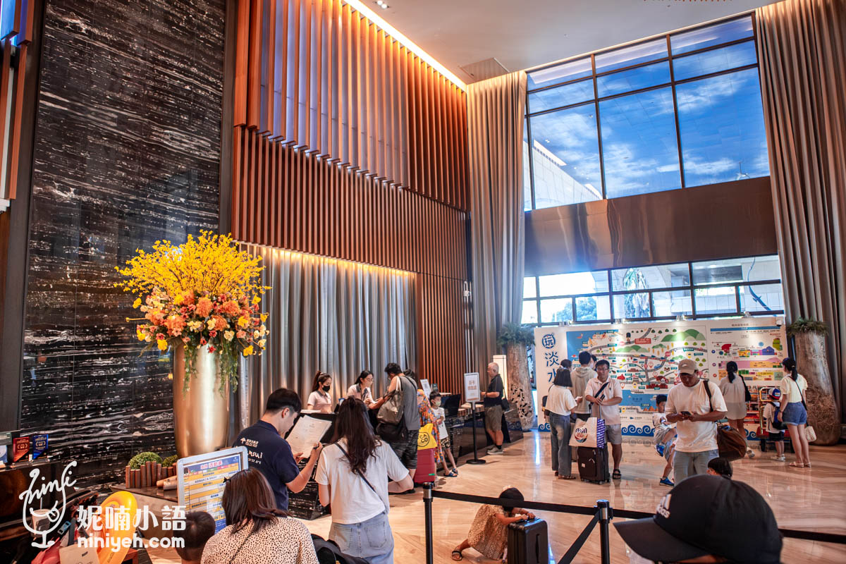 這張照片展現了一個現代化酒店大堂的繁忙景象，透過高聳的垂直木條格柵與大理石牆面，營造出一種奢華且宏偉的空間感。大廳內旅客穿梭其中，有人在櫃檯辦理手續，有人則圍繞在色彩鮮艷的景點導覽圖前，顯見該場所具備強烈的觀光服務功能。角落裡裝飾著巨大的盛開花藝，與自然採光交織出熱情款待的氛圍，精準捕捉了旅行中充滿活力的起點時刻。