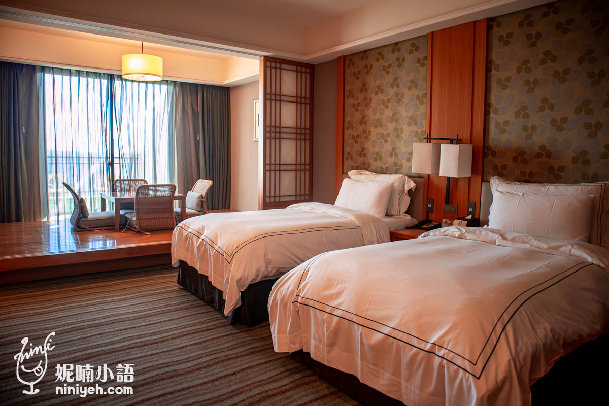 這張照片展示了一個融合現代舒適與日式禪意的飯店客房，呈現出寧靜且高雅的休憩空間。室內配置了兩張整潔的單人床，並以木質壁板與和風花紋牆面營造出溫暖的視覺層次。後方則設有一個架高式的和室區域，透過推拉門與採光充足的落地窗，將室內與戶外自然景觀巧妙連結。整體的設計旨在提供旅客一個能放鬆身心的精緻環境，展現出對空間美學與居住品質的細膩追求。