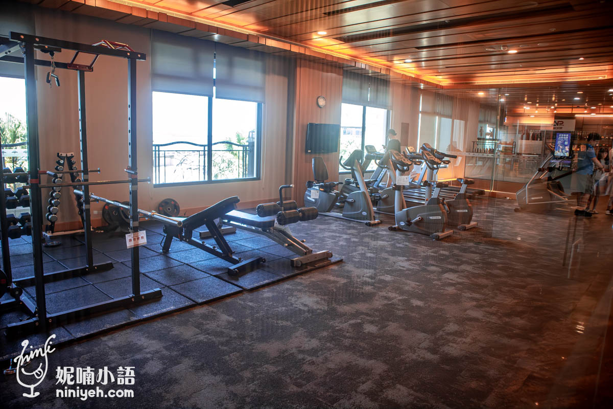 這張照片展示了一個設計精良且設施齊全的健身空間，旨在為使用者提供多樣化的運動選擇。室內佈局將重量訓練區與有氧運動器材（如跑步機和健身車）有序地分開，並透過大片窗戶引入自然光，營造出明亮且舒適的運動環境。整體裝潢採用溫暖的木質天花板與深色地墊，這種設計不僅兼顧了功能性與美學平衡，更展現出一種專業且具現代感的健身氛圍。