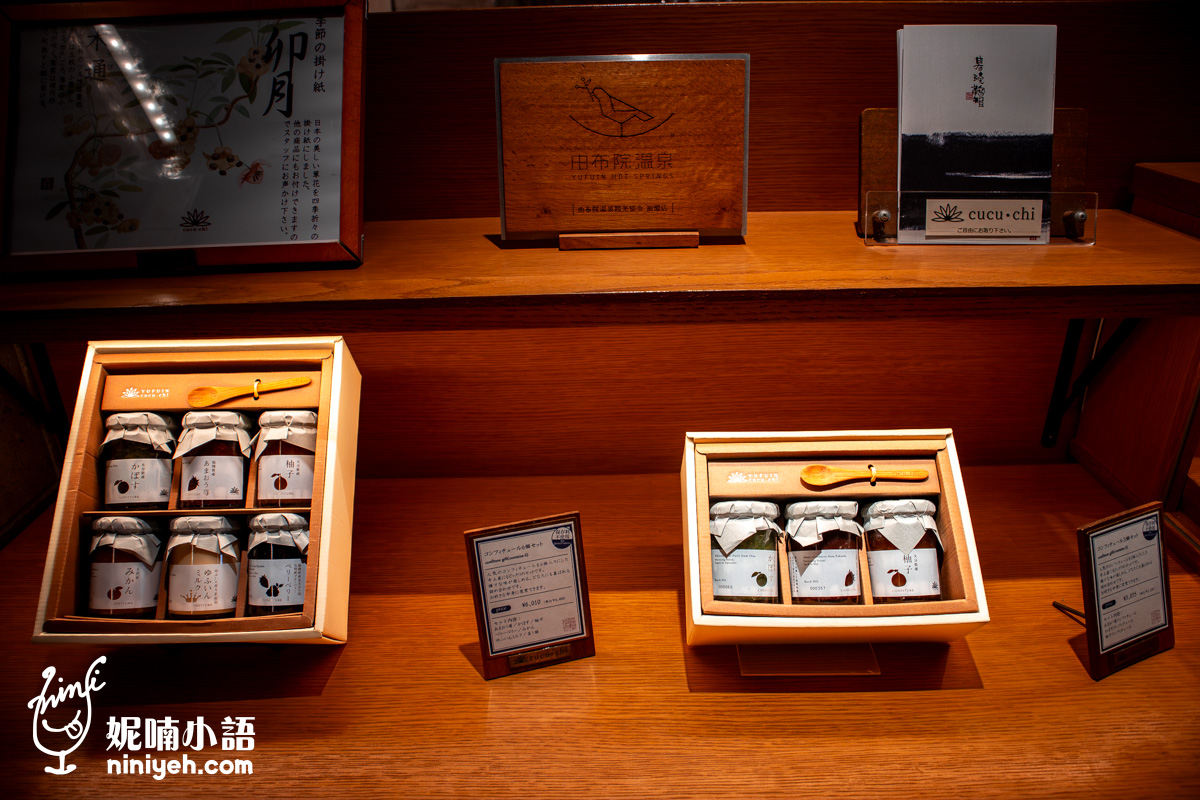 由布院鞠智（cucuchi）店內展示的手工果醬禮盒。木質層架上擺放著三入與六入裝的精緻果醬禮盒，玻璃罐上方附有木製小湯匙，上方層架則陳列「由布院溫泉」木製紀念牌與店鋪簡介摺頁，整體氛圍呈現日式文青與優雅伴手禮風格。