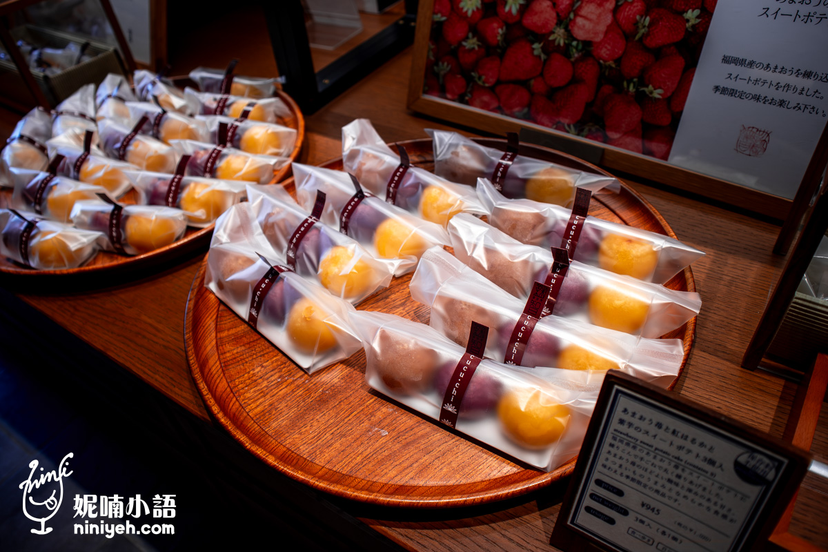 由布院鞠智（cucuchi）店內販售的季節限定點心組合。圓形木托盤上擺放著三入一組的透明長條包裝，內含草莓、紫薯與紅春香地瓜三種顏色的迷你地瓜燒（Sweet Potato Cake），前方設有標示售價 ¥945 的日文介紹牌。