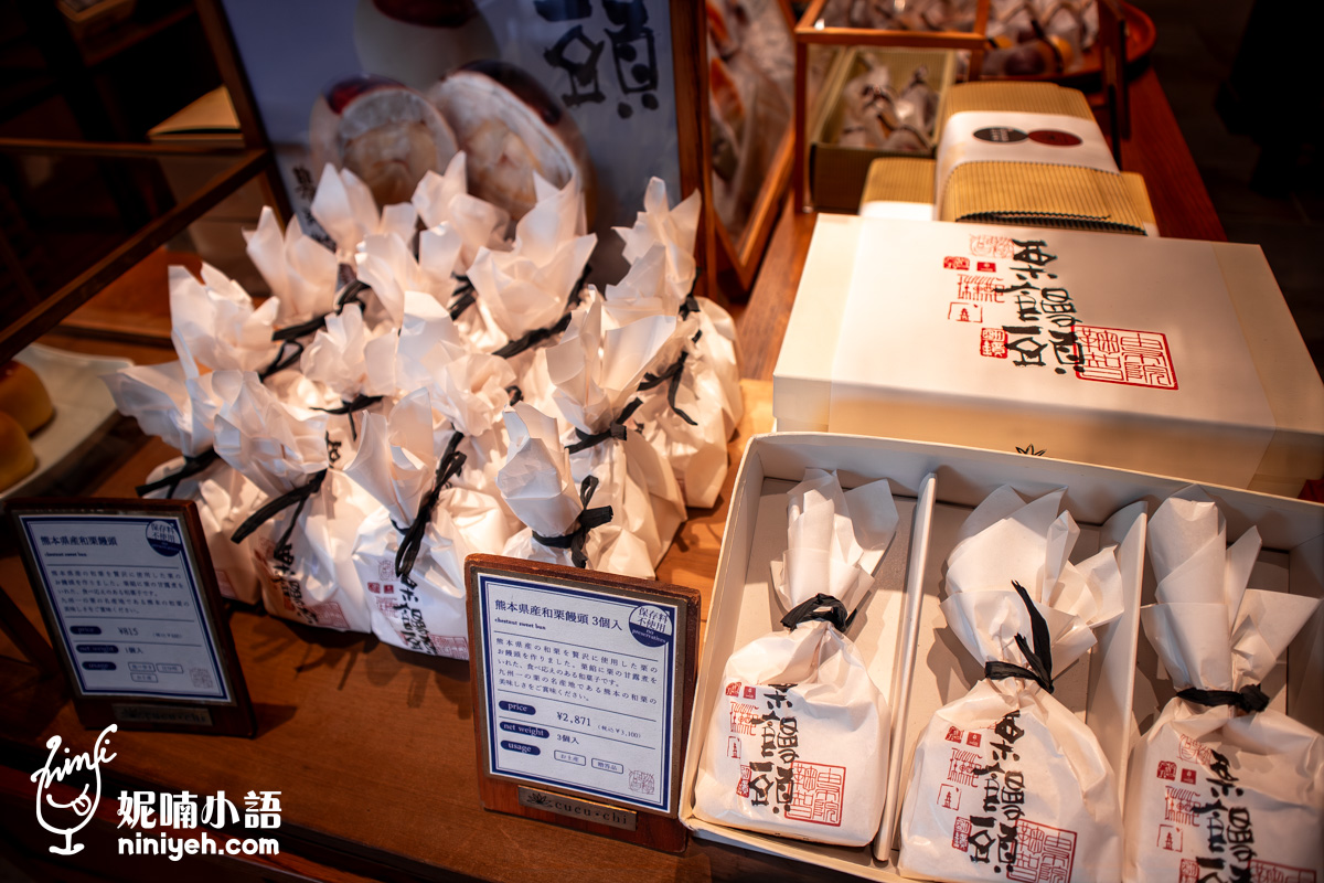 由布院鞠智（cucuchi）店內販售的「熊本縣產和栗饅頭」伴手禮。木檯上陳列著以白色和紙、黑色細繩精緻包裝的單個栗子饅頭點心，右側則有三入裝的禮盒與精美包裝外盒，前方設有日文產品說明的木質介紹牌。