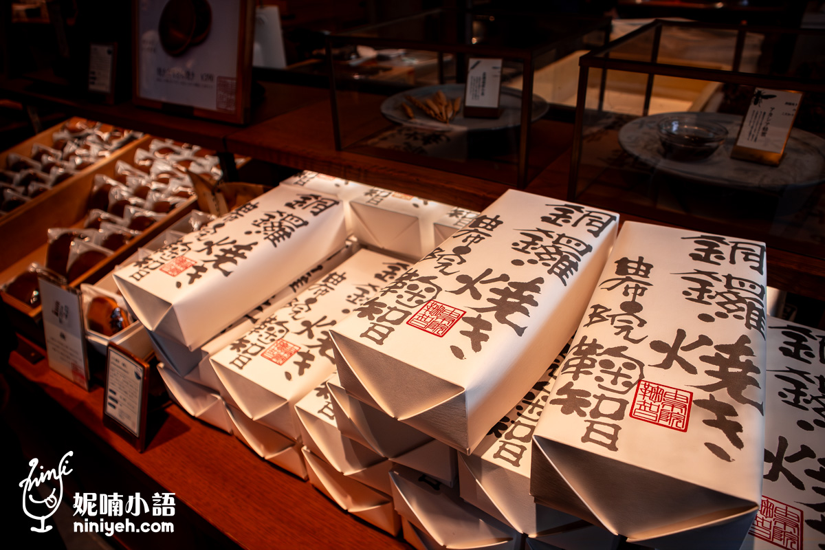 由布院鞠智（cucuchi）店內陳列的銅鑼燒伴手禮盒。白色包裝紙上印有黑色的「銅鑼焼き」與「由布院鞠智」日文書法字體，並蓋有紅色印章，整齊堆疊在木質展示檯上。左側背景還能看到單個包裝的裸裝銅鑼燒，展現出精緻且具傳統日式質感的和菓子店鋪氛圍。