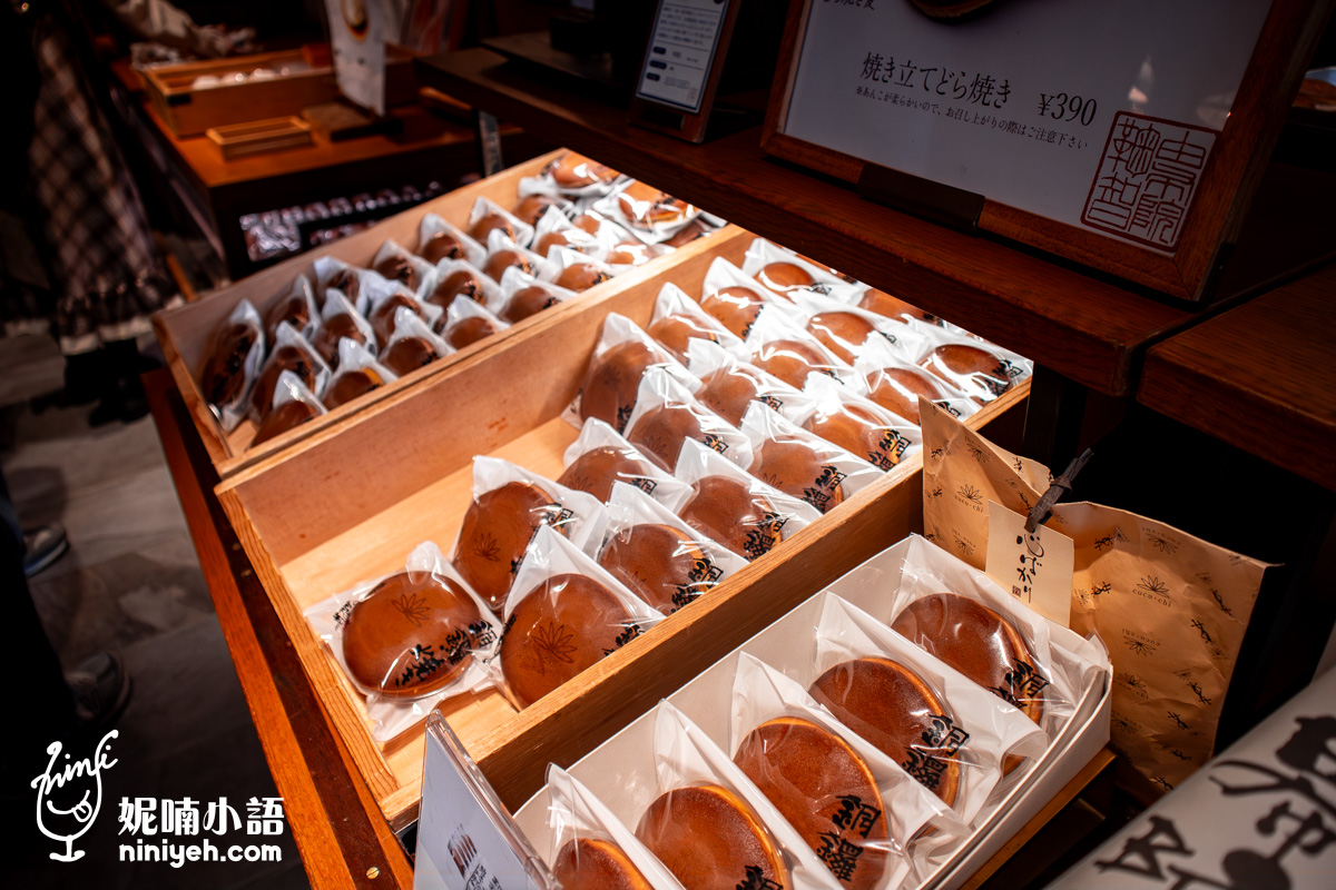 由布院鞠智（cucuchi）店內木質展示檯上陳列的單個獨立包裝銅鑼燒。透明塑膠包裝內透出金黃油亮的銅鑼燒餅皮，上方設有售價標籤標示「焼き立てどら焼き ¥390」，右側則有精緻的紙袋包裝，呈現傳統日式和菓子店溫馨典雅的伴手禮販售場景。