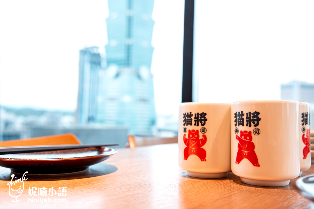 遠百信義 A13 貓將日本料理的餐桌視角,擺放著印有可愛紅色貓咪圖騰的專屬茶杯與日式餐具。在這裡用餐不僅能品嚐美食,還能透過玻璃窗欣賞迷人的台北 101 高空城市景觀,享受極具質感的用餐氛圍