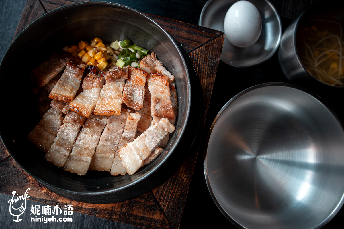 「金美麗人」招牌海鹽烤五花肉釜飯定食的俯瞰特寫,鐵鍋內鋪滿切得厚實、表皮金黃焦香的現烤豬五花肉片,旁邊配有黃澄澄的玉米粒與新鮮蔥花,並附上空碗、豆芽高湯和一顆生雞蛋。