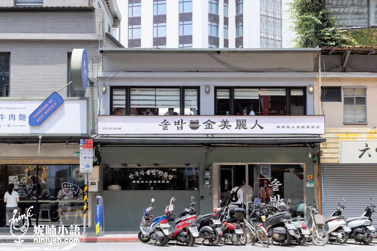 位於台北士林捷運站附近的「金美麗人 韓國直火釜飯專賣店」兩層樓灰色系店面外觀,招牌帶有韓文字體,一樓為大片落地窗與候位區,門口停放多輛機車。