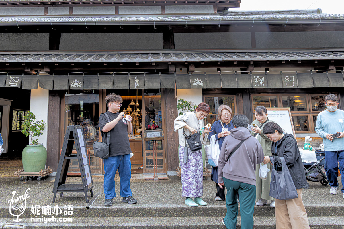 由布院人氣美食「鞠智（cucuchi）」店門口遊客聚集的熱鬧場景。多位旅客站在古樸的雙層日式黑瓦建築前，手裡拿著剛買到的現做銅鑼燒現場品嚐。屋簷下掛著印有蓮花標誌與「由布院鞠智」字樣的深色暖簾，展現湯之坪街道熱門伴手禮店的日常街景。