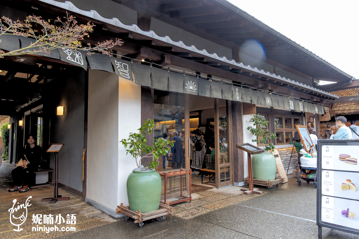 由布院湯之坪街道上的「鞠智（cucuchi）」和菓子專賣店全景。古樸的日式黑瓦屋頂與白色牆面搭配木質大門，屋簷下掛滿一整排深色日式暖簾。門口立有展示銅鑼燒與點心圖片的白色告示牌，並擺放大型綠色陶甕盆栽點綴，展現洗練古樸的民宅民風。