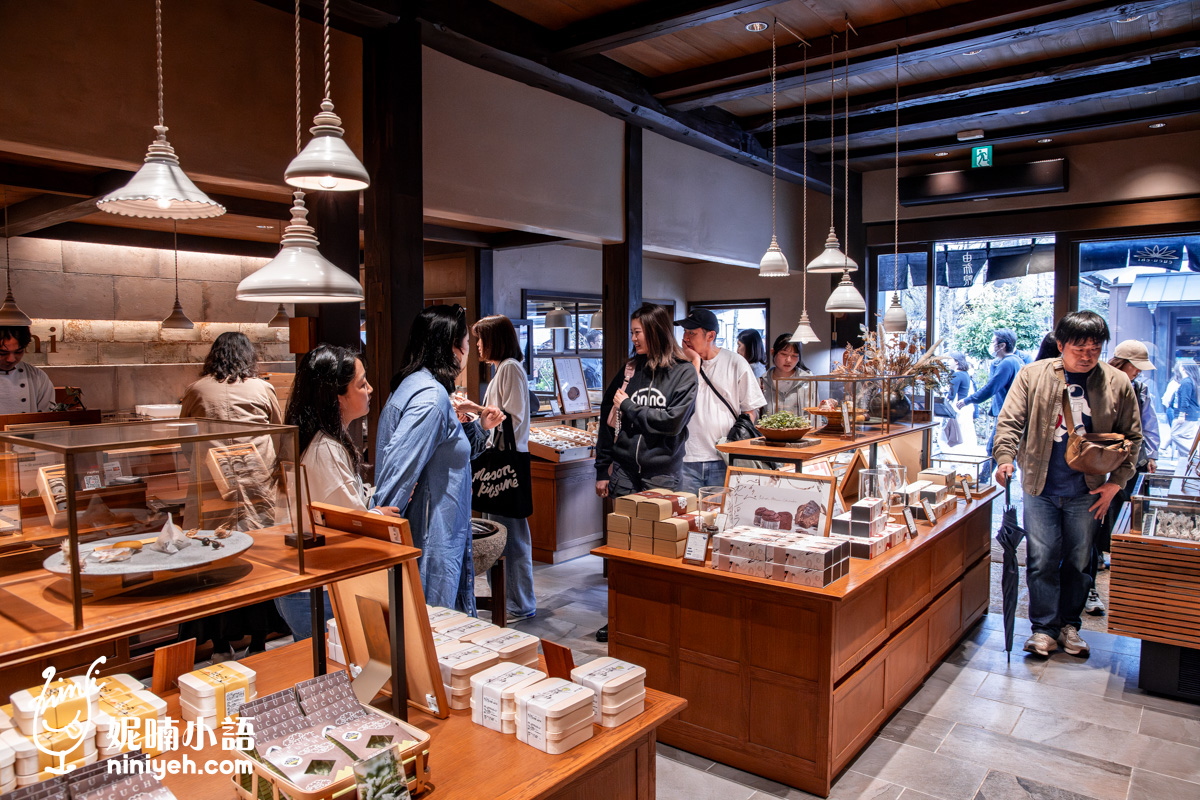由布院鞠智（cucuchi）店內熱鬧的購物景象。日式傳統木造建築內擁有高挑的木樑屋頂與復古典雅的白色吊燈，多張木質展示檯上陳列著精緻的和菓子伴手禮，許多遊客正在店內慢逛、挑選商品，採光良好的大門口可見戶外熱鬧的湯之坪街道。