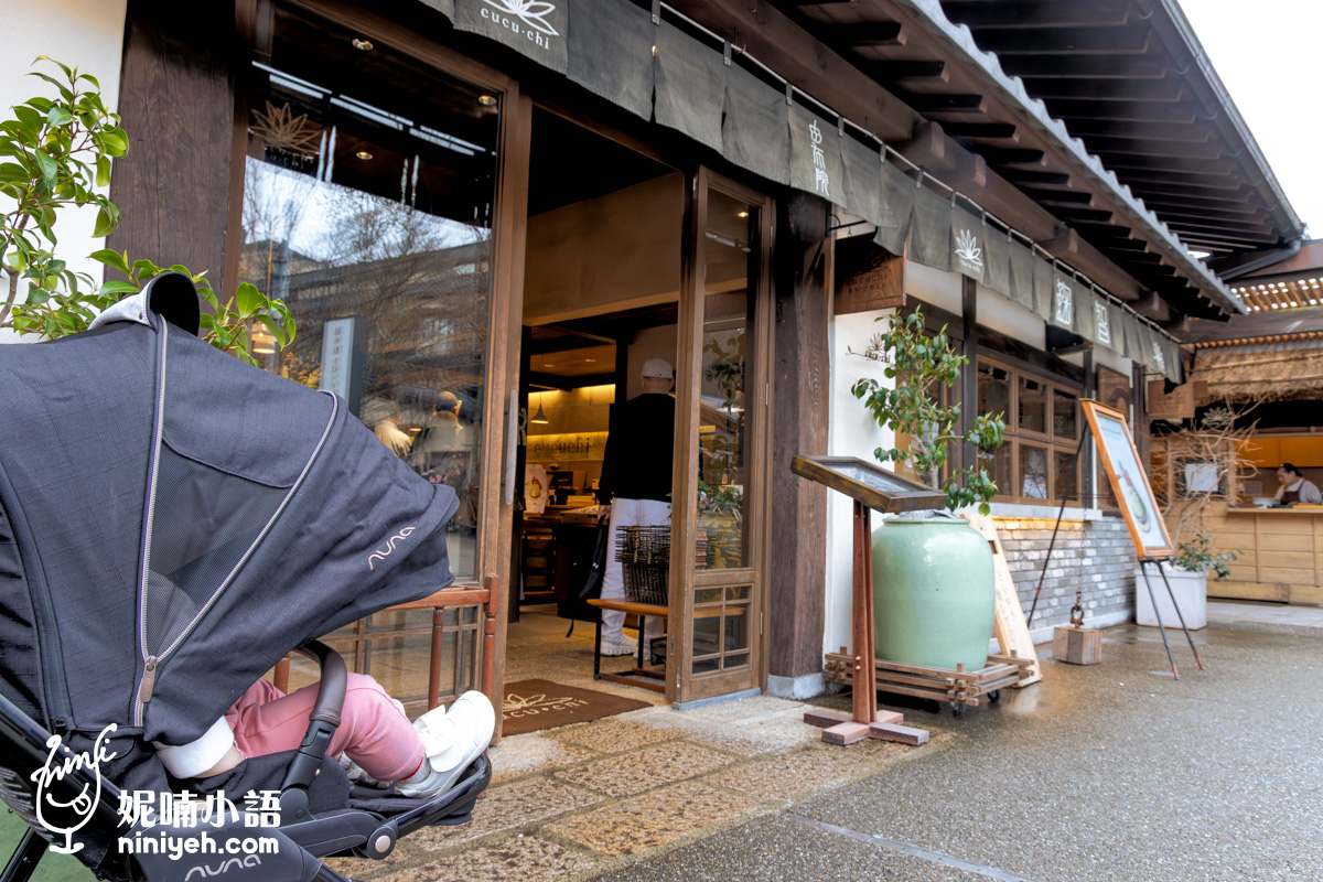 女兒的嬰兒車停在由布院「鞠智（cucuchi）」店門口。木造的日式傳統建築外觀掛有印著「由布院」與「鞠智」的深色暖簾，門口擺放著大型綠色陶甕植栽，透過玻璃門可以看見店內溫馨的燈光與選購商品的遊客。