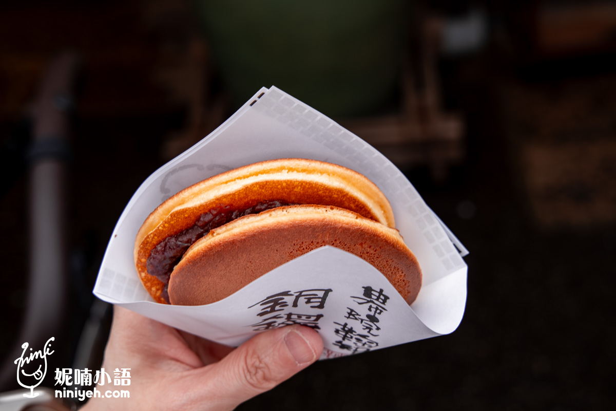 由布院名產「鞠智」銅鑼燒（由布院鞠智銅鑼焼），外皮鬆軟金黃、紅豆內餡飽滿，以白色和紙包裝，上面印有「銅鑼燒」、「由布院鞠智」等日文字樣，由人手持著，背景為日式傳統氛圍。