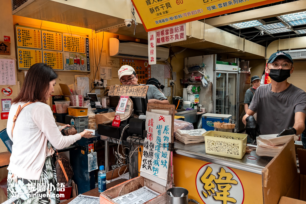 台北小巨蛋美食緯大雞排點餐與取餐櫃檯實拍。顧客正在前台交單結帳，戴著口罩與手套的小老闆正忙碌地將打包好的炸雞排遞送給客人，櫃檯上放有「請先寫單再排隊」的告示牌。