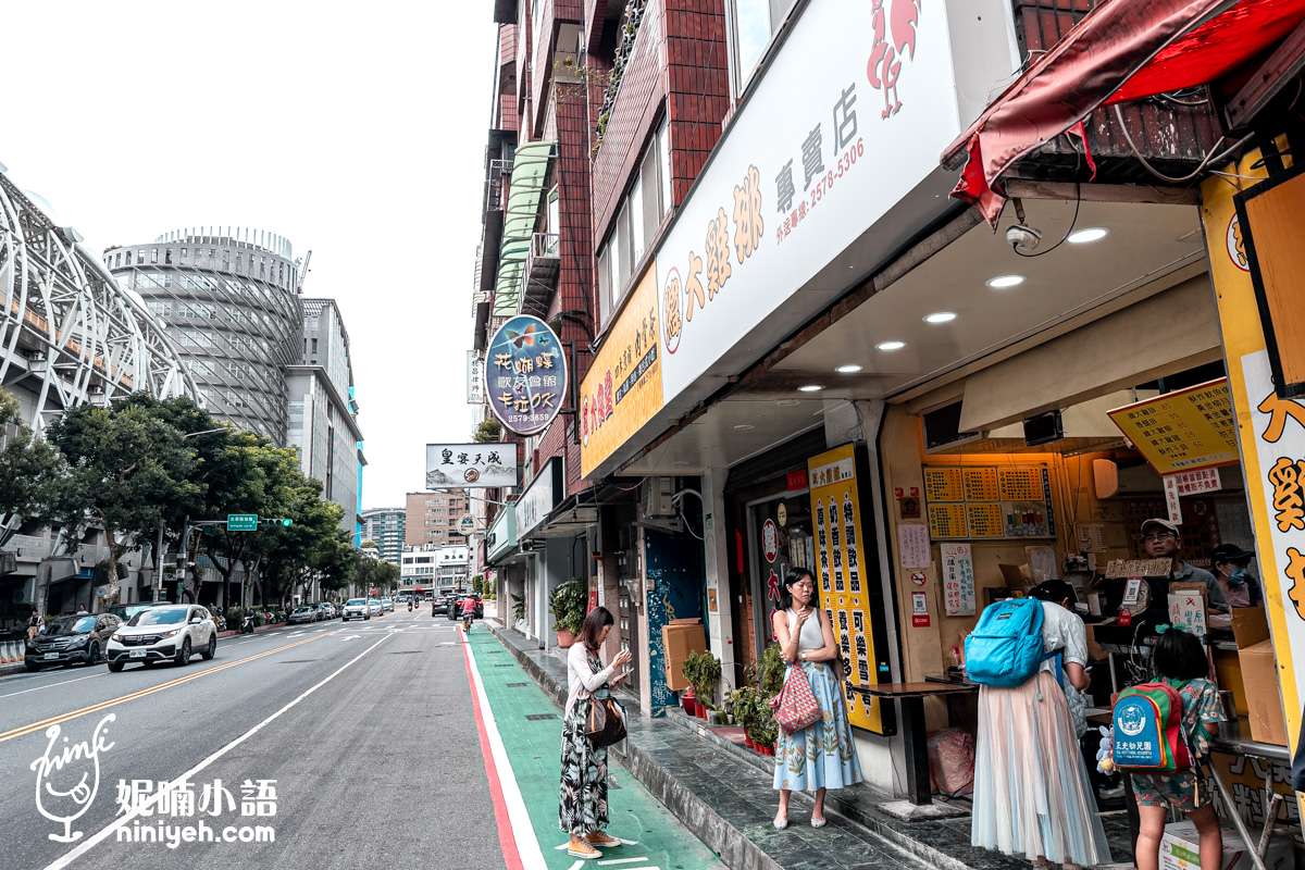 台北小巨蛋旁北寧街景上的緯大雞排專賣店實景。黃色招牌和詳細菜單看板清晰。一個背著藍色書包的孩子和一個成年人正在櫃檯前等待。兩名女性站在人行道上，左側道路可見台北小巨蛋的現代化結構和許多其他招牌