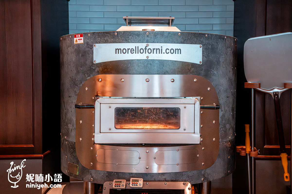 餐廳披薩烘焙坊內設置的 Morello Forni 義大利頂級手工窯爐,圓柱形爐身帶有工業風金屬質感與鉚釘裝飾,中央設有透明耐熱玻璃的烘烤艙門。右側擺放著烘烤披薩專用的不銹鋼長柄披薩鏟,前方配有數位計時器。