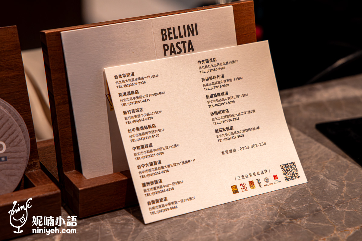 櫃檯木質名片架上展示的 BELLINI PASTA 品牌資訊卡特寫。白色卡片上詳細列出包含台北車站店、新莊宏匯店、南港潤泰店等全台多間分店的地址與電話,右下方印有三商企業餐飲品牌標誌與客服專線。