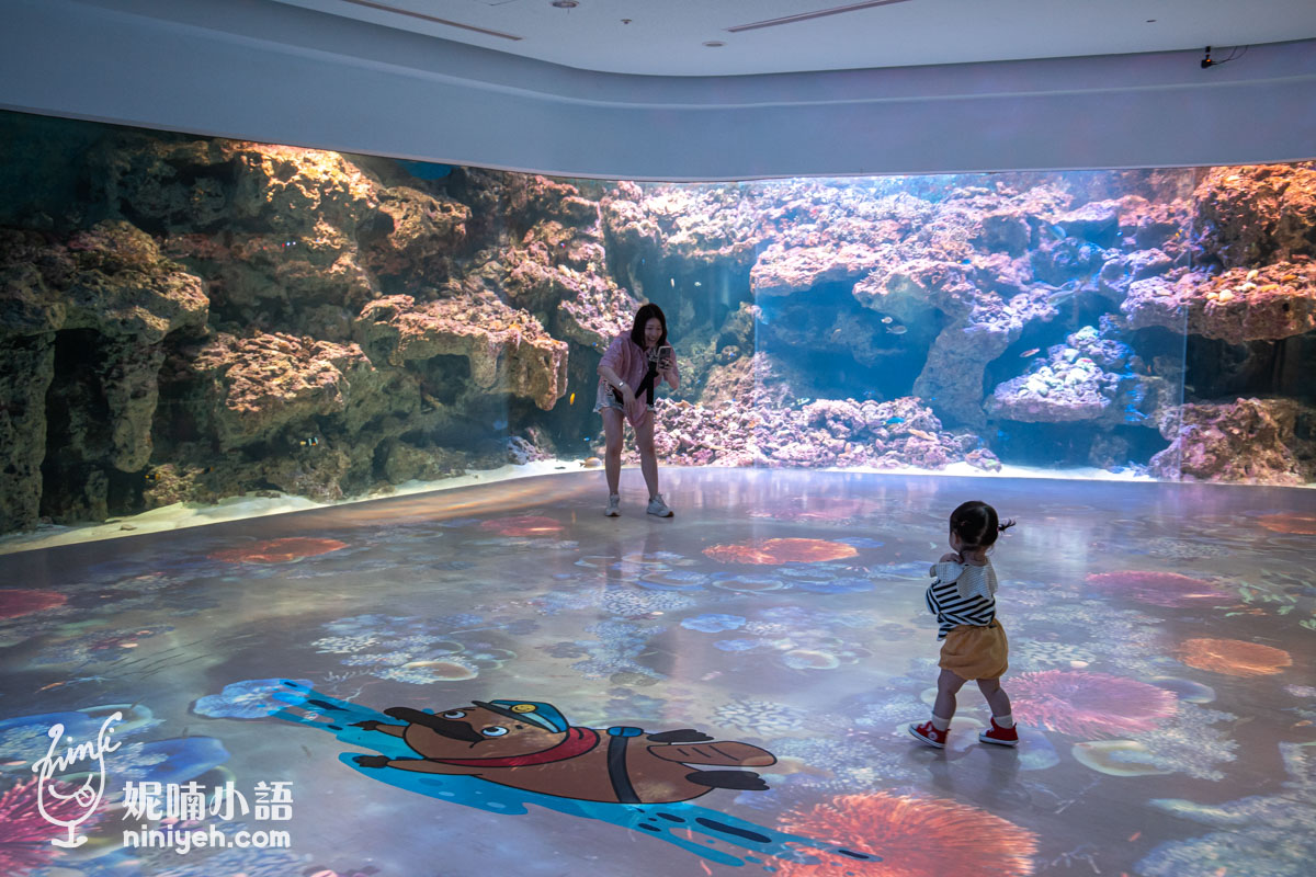 我們正在 XPARK 水族館的互動地板上,微笑著捕捉女兒的身影,女兒正好奇地看著地板上的珊瑚和卡通河狸投影。身後的巨大珊瑚礁魚缸閃閃發光,裡面有許多魚兒在游動。